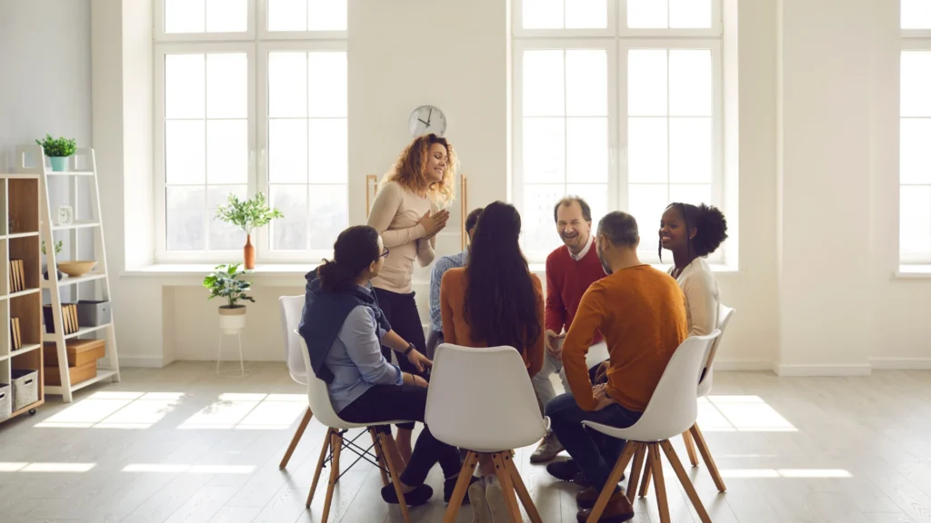 Group therapy session in a bright counseling space, with participants seated in a circle listening and sharing during a supportive mental health discussion.