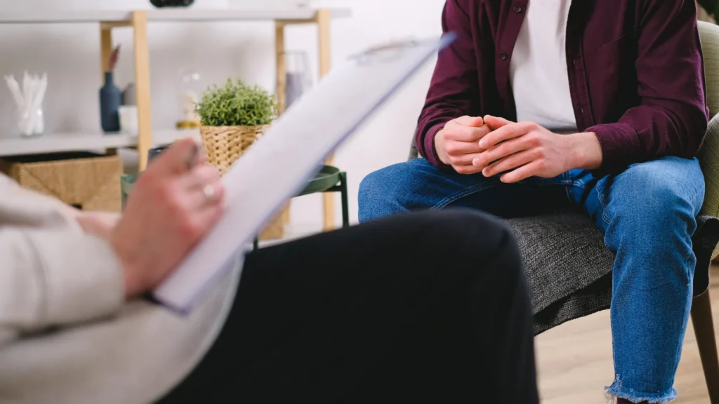Person seated in a therapy session speaking with a mental health professional, with the therapist holding a clipboard in the foreground and a calm office setting in the background.
