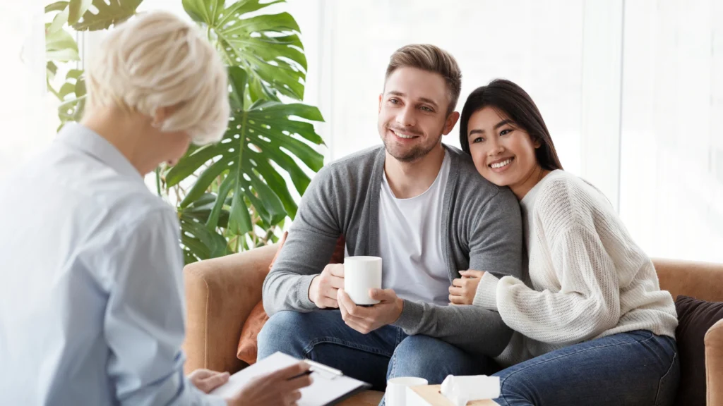 Middle-aged couple sitting together on a couch at home, smiling and raising their arms to form a heart shape, representing connection, support, and relationship wellness.